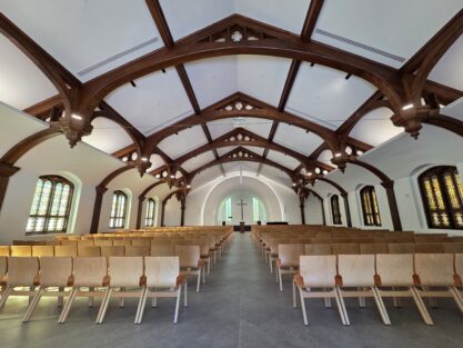 Empty chapel with rows of wooden chairs facing a cross at the front, under a vaulted ceiling with wooden beams and stained glass windows.