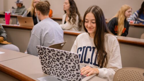 Girl sitting in a classroom full of students, typing on her laptop.