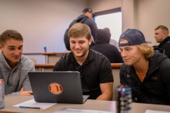 3 students sitting in a classroom looking at 1 laptop.