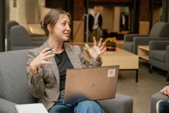 girl sitting in a chair explaining something, while resting a laptop on her lap.
