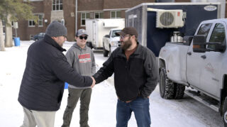 Two men in warm winter clothes shake hands in front of another man while a third man stands just past them. There is a truck towing a trailer, and the truck has