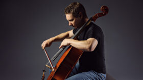 A man in a black tee shirt and jeans plays a cello.