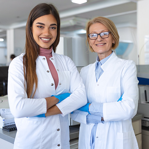 Two women in white lab coats stand with their arms crossed in a laboratory. 