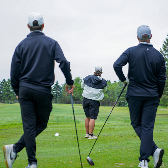 Two golfers in blue outfits and white hats hold clubs while watching a fellow golfter tee off. 