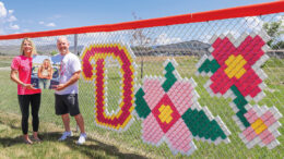 Two people stand by a decoration on a chain-link fence depicting a letter 