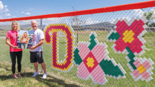 Two people stand by a decoration on a chain-link fence depicting a letter 