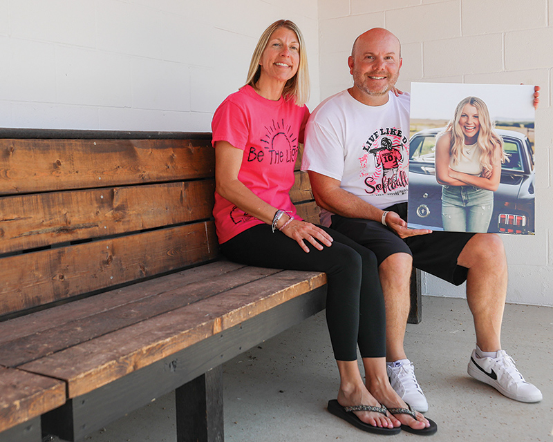 Two people sit on a wooden bench holding a portrait of their teenage daughter.
