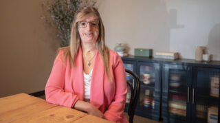 A woman wearing a pink blazer and a white shirt sitting at a table with a bookcase behind her.