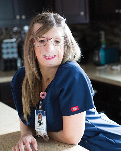 A woman wears blue nursing scrubs and her employee ID. 