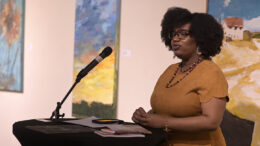 A woman stands speaking at a microphone in front of a few paintings.