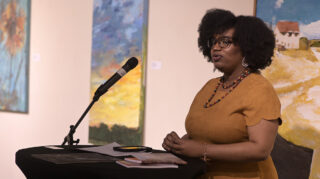 A woman stands speaking at a microphone in front of a few paintings.