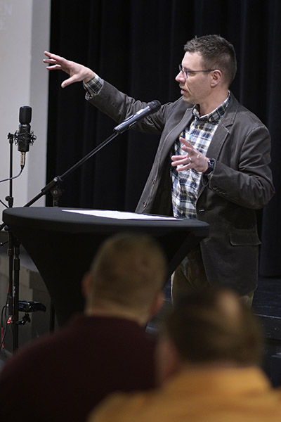 A man guestures with his hands while speaking into a microphone in front of an audience.