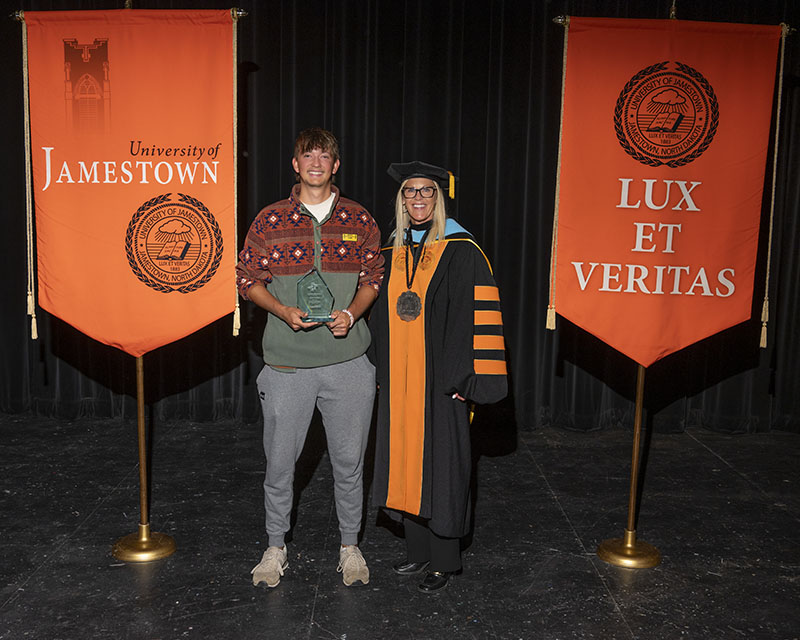 A man holds a glass trophy while standing next to a woman in academic regalia. There are banners next to them that read "University of Jamestown" and "LUX ET VERITAS."