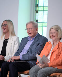 From left, a woman with blonde hair and wearing black glasses and a white blazer, a man in a grey suit, and a woman in an orange jacket laugh together. There is a green stained glass window behind them.