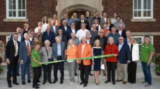 Several dozen people stand on the steps in front of a red brick chapel. The front people hold a green ribbon with two in the middle holding giant scissors to cut the ribbon.