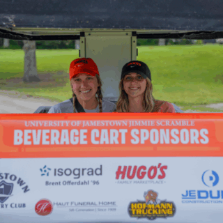 Two University of Jamestown Jimmie Scramble beverage cart drivers smile behind a sign listing event sponsors.