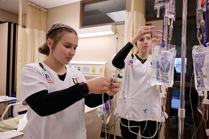 Two women in nursing scrubs hold tubing and connect it to saline solution bags. 