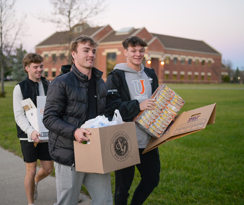 Four men carry boxes of food and supplies across a college campus.
