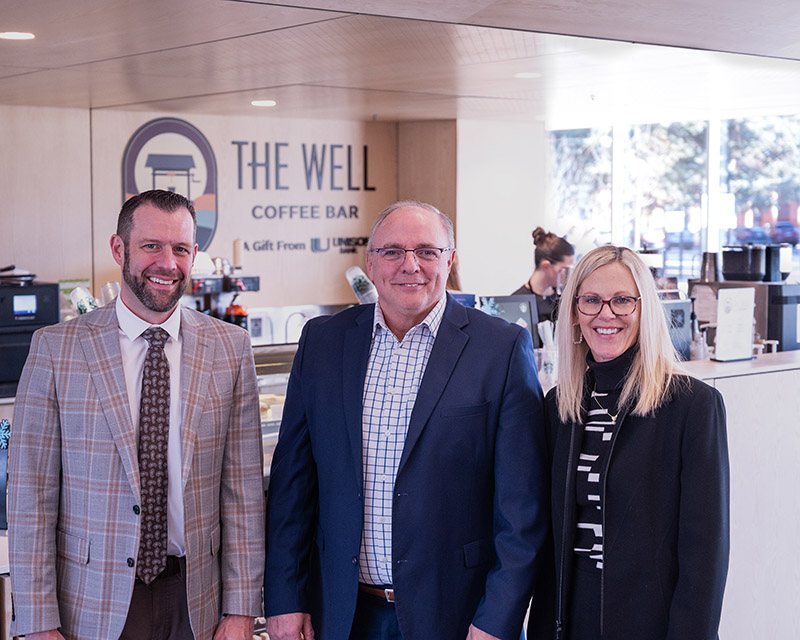 Three people stand and smile in front of a coffee shop kitchen. The words "The Well" are visible on the wall behind them.