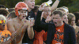 A man in a blazer and T-shirt walks past a group of cheering students on a football sideline. The man pumps his arm with a water bottle in his hand.