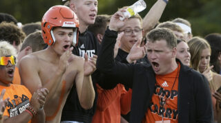 A man in a blazer and T-shirt walks past a group of cheering students on a football sideline. The man pumps his arm with a water bottle in his hand.