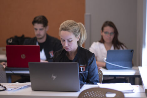 Three university students work on laptops and tablets in a classroom, focusing on their studies. The student in the foreground wears a 