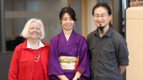 Three people smiling and standing together indoors. The person in the middle is wearing a purple kimono with a decorative obi, while the person on the left is dressed in a red outfit, and the person on the right is wearing a gray button-up shirt.