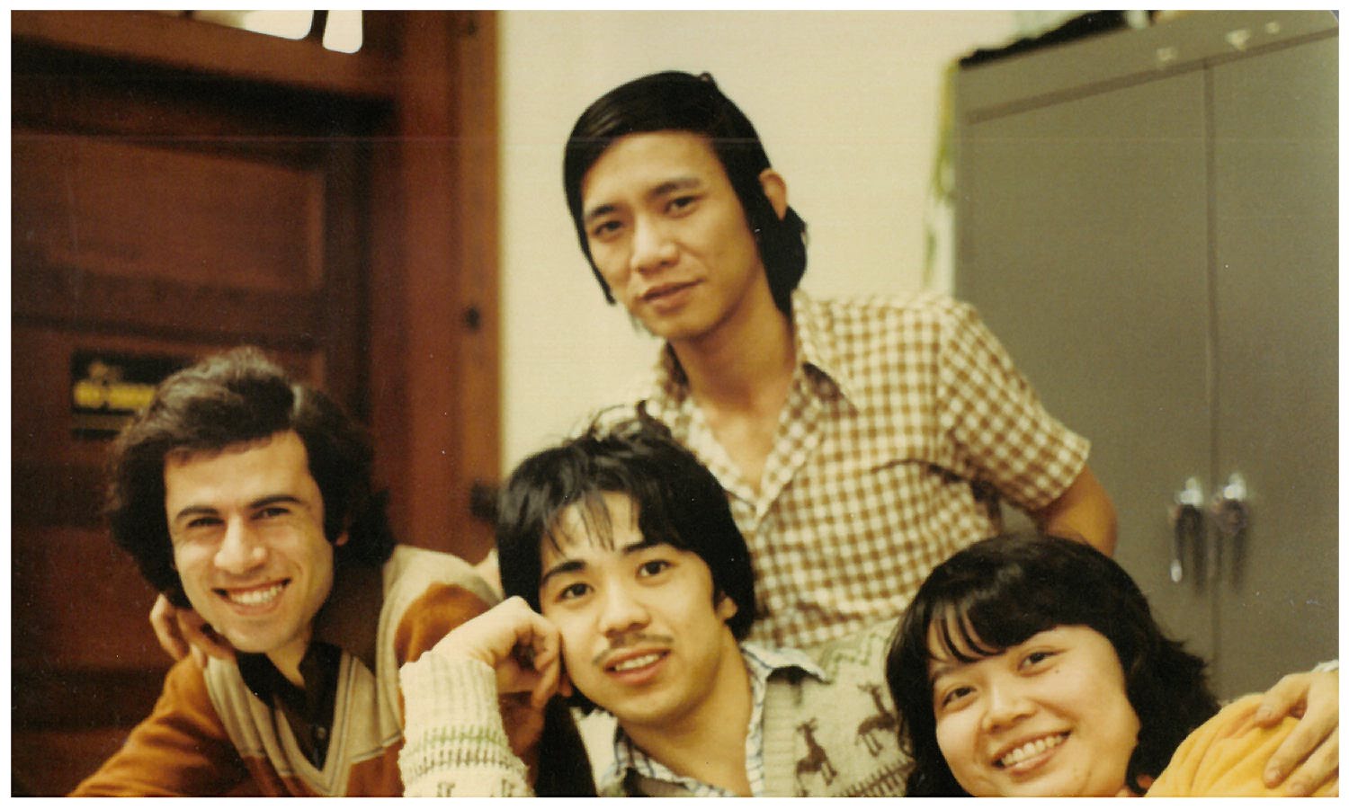 A group of four people smiling and posing together indoors. They appear to be in a casual setting with a wooden door and a cabinet in the background.