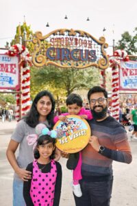 A family of four poses in front of the Storybook Circus entrance at Walt Disney World, holding a bright yellow "1st Visit!" sign featuring Mickey Mouse and friends. The family is smiling, and the scene is decorated with festive holiday garlands.