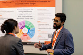 A man wearing glasses and a blazer discusses his research with a woman in a suit in front of a poster titled 