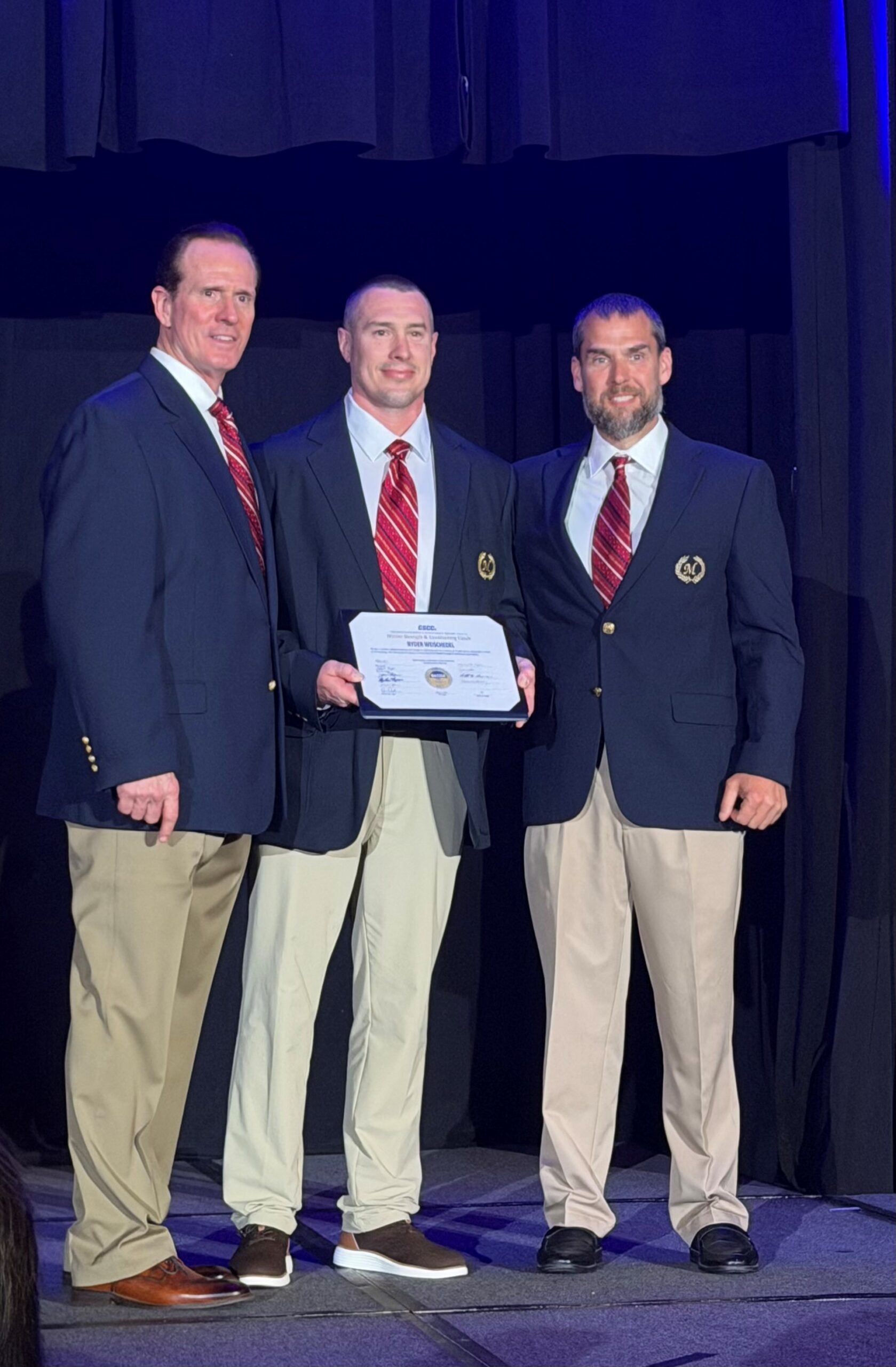Three men pictured together on stage. The individual in the center is holding an award.