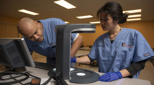male and female in scrubs look through microscope in classroom
